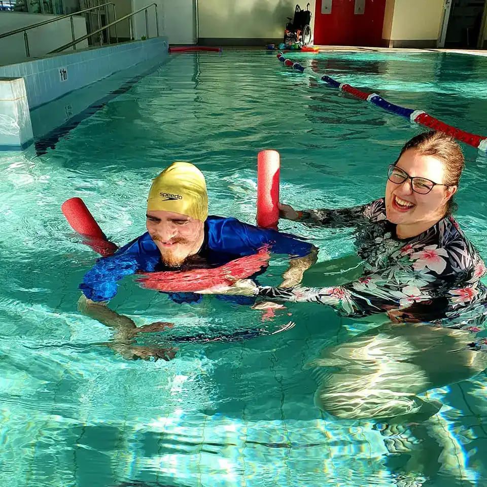 An image of physiotherapist, Heather, and one of her clients in the pool. The client, a young man wearing a yellow swim cap and blue rashie, is paddling along with a red pool noodle under his arms and a smile on his face.